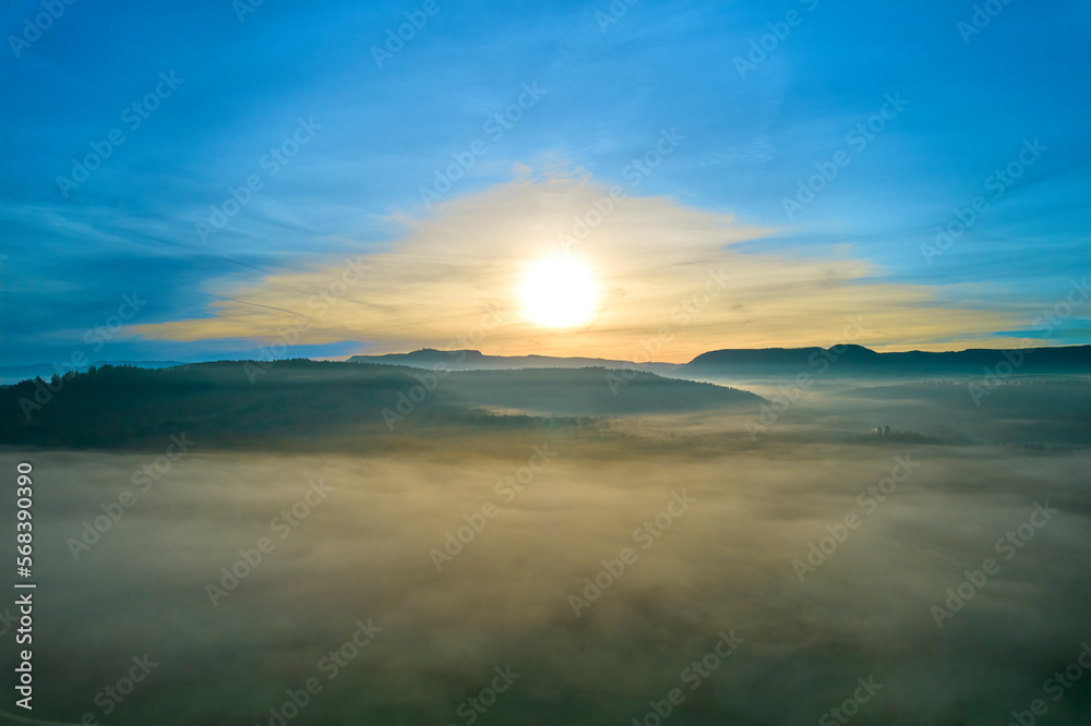 Aerial view of foggy landscape at sunrise. Mountains in front of valley with yellow sun in autumn. Germany, Nurtingen, Tiefenbachtal.