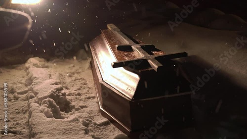 A closed coffin lies on the ground in the snow at night. Strong blizzard. Burning car headlights