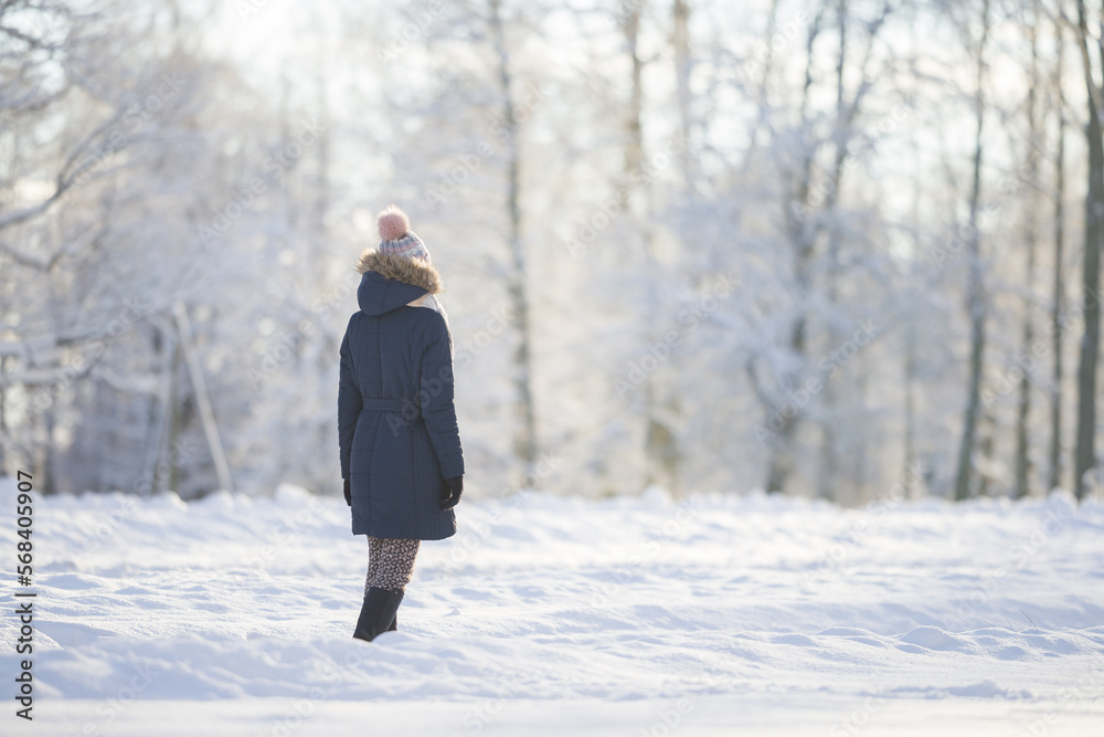 Young adult woman slowly walking on white snow covered sidewalk at park in cold sunny winter day. Spending time alone. Enjoying peaceful stroll. Back view. Young adult woman slowly walking on white snow covered sidewalk at park in cold sunny winter day. Spending time alone. Enjoying peaceful stroll. Back view.