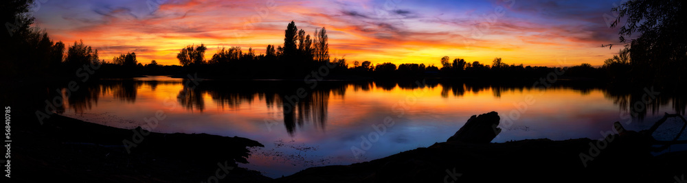 Magnificent colorful sunset at a lake, extra wide panorama with dark silhouettes of trees and the lakeshore, with the dramatic pretty sky reflected in the water