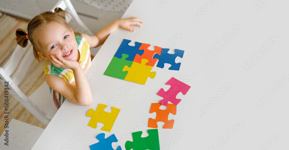 A cute beautiful little innocent girl is sitting at a table with large colorful puzzles. A puzzle for preschool development. The symbol of autism A cute beautiful little innocent girl is sitting at a table with large colorful puzzles. A puzzle for preschool development. The symbol of autism