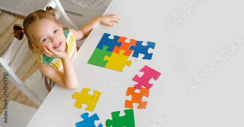 A cute beautiful little innocent girl is sitting at a table with large colorful puzzles. A puzzle for preschool development. The symbol of autism