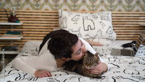 Beautiful woman is relaxing on the bed with her cat. The woman is lying down and playing with the gray cat.