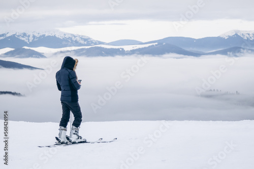 A snowboarder in a red jacket stands on top of a mountain and looks into the distance