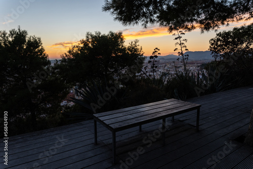 Von Pflanzen umgebene zugewachsene leere Parkbank ohne Lehne mit Aussicht auf eine spanische Stadt bei farbenfrohem Sonnenuntergang 