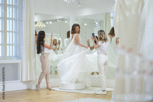 Women taking photographs of a female friend trying on wedding dress. Women in wedding dress fitting room.