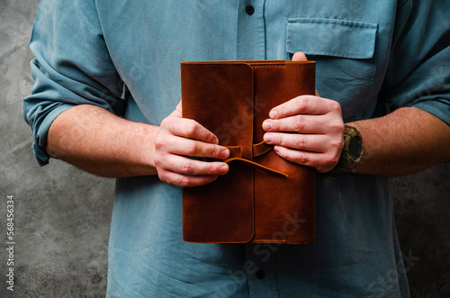 A man in a shirt holds a leather notebook in his hands against a concrete wall. Lifestyle.