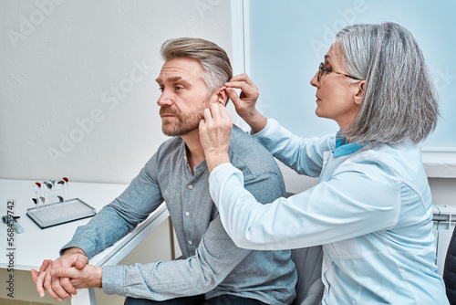 Fototapete Experienced female doctor fitting Intra-The-Ear hearing aid into patient's ear while consultation in audiology clinic
