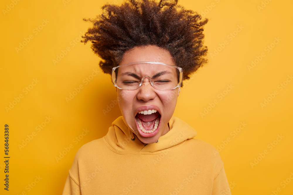 Wild crazy teenage girl dressed in hoodie wears big transparent eyeglasses shouts loudly reacts to something terrifying isolated over vivid yellow background. Negative emotions and feelings concept Wild crazy teenage girl dressed in hoodie wears big transparent eyeglasses shouts loudly reacts to something terrifying isolated over vivid yellow background. Negative emotions and feelings concept