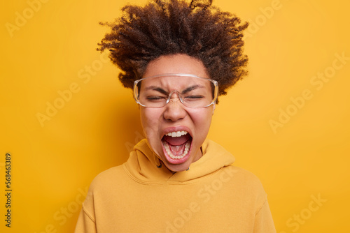 Wild crazy teenage girl dressed in hoodie wears big transparent eyeglasses shouts loudly reacts to something terrifying isolated over vivid yellow background. Negative emotions and feelings concept