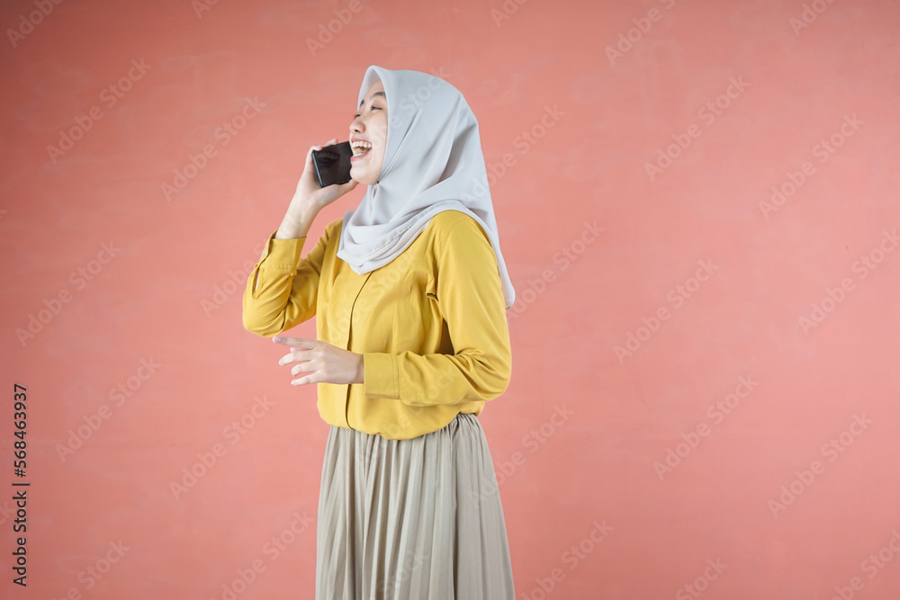 Obraz premium Beautiful Asian woman in yellow shirt and hijab smilling cheerfully holding smartphone on brown background 