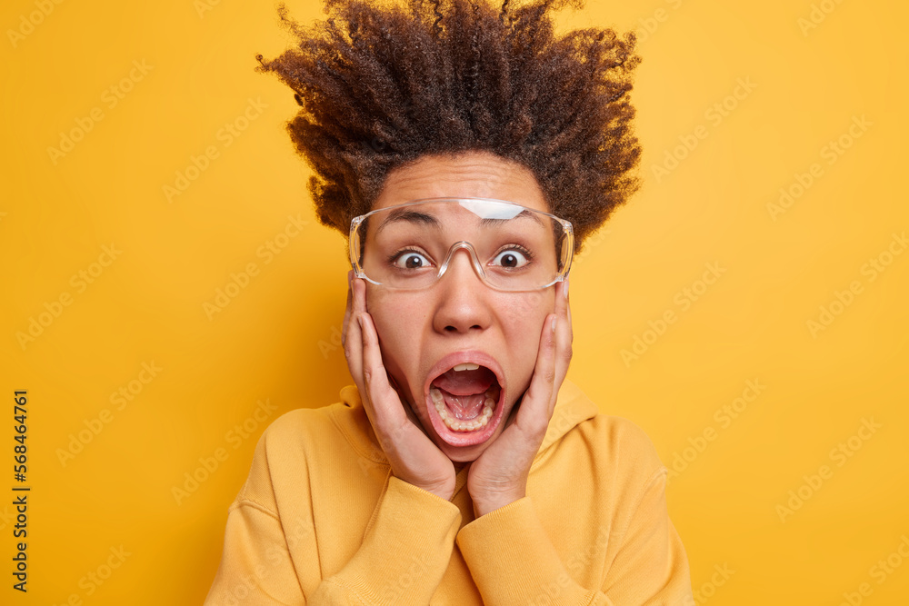 Crazy emotional young woman with curly hair standing up keeps hands on cheeks exclaims loudly keeps mouth widely opened wears transparent eyeglasses and hoodie isolated over yellow background Crazy emotional young woman with curly hair standing up keeps hands on cheeks exclaims loudly keeps mouth widely opened wears transparent eyeglasses and hoodie isolated over yellow background