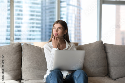 Free Download Thoughtful serious millennial freelancer girl working on online project on home couch, holding laptop, touching chin, looking away, up with doubts, thinking over ideas, planning tasks - GFXLoad