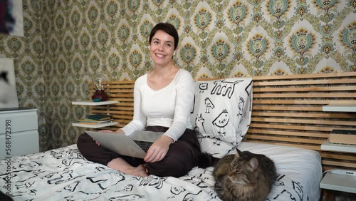 Beautiful woman is working on the computer while listening to music on headphones. The woman is sitting on the bed and petting her cat.