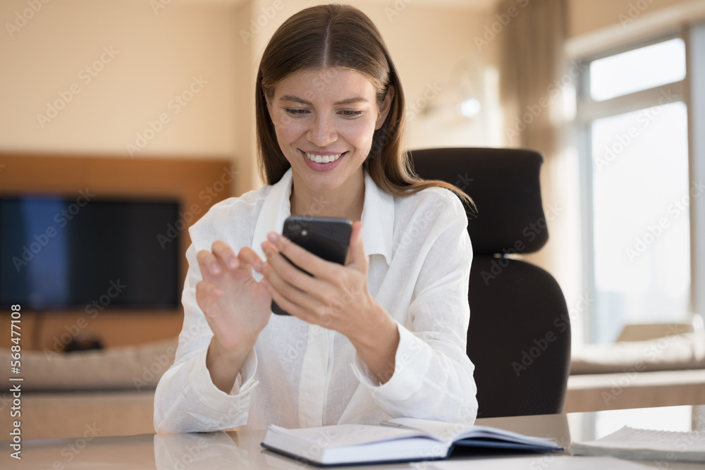 Happy engaged young businesswoman using mobile phone for Internet communication, sitting at notebook on work table at home office workplace, chatting online, typing Happy engaged young businesswoman using mobile phone for Internet communication, sitting at notebook on work table at home office workplace, chatting online, typing