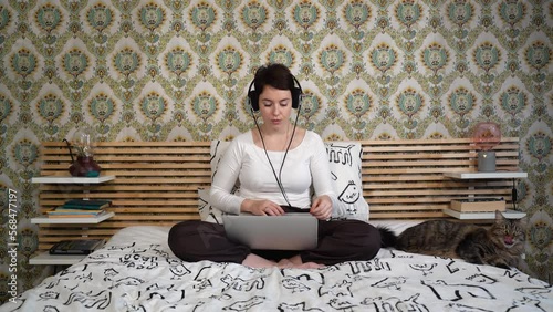 Beautiful woman is working on the computer while listening to music on headphones. The woman is sitting on the bed and petting her cat.