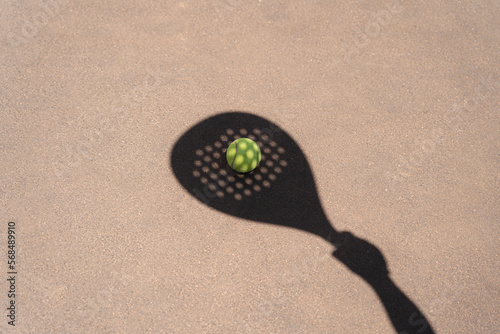High angle of yellow ball placed on court and covered with shadow of crop person holding padel racket