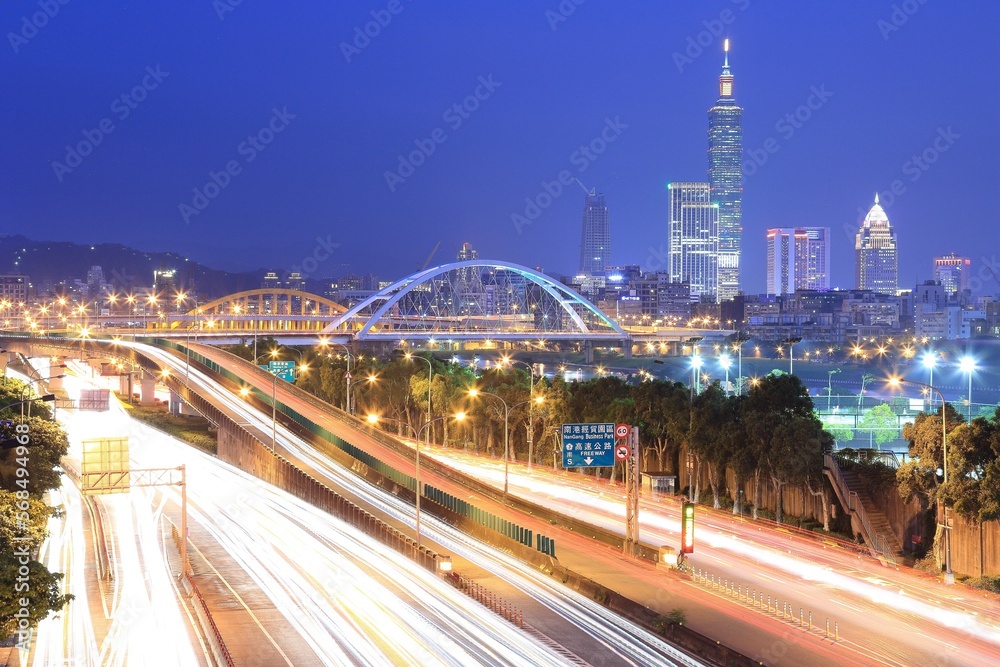 Naklejka premium Night scenery of Taipei City with view of Taipei 101 Tower in downtown, MacArthur Bridge across river and car trails on Dike Avenue ~ Taipei cityscape at dusk by riverside (long exposure effect)
