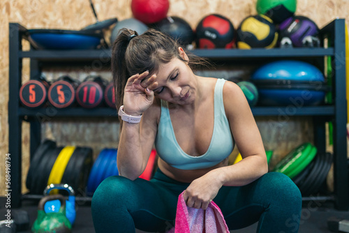 Young sporty woman in the gym sitting on top of a soft wall ball holding a hand to her forehead with signs of fatigue and suffering. photo