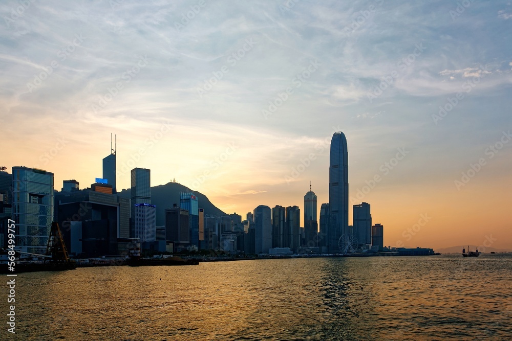 Sunset scenery of Hong Kong under dramatic sky, with famous landmark International Finance Center (IFC) standing among skyscrapers by Victoria Harbour & the setting sun with golden glow in background