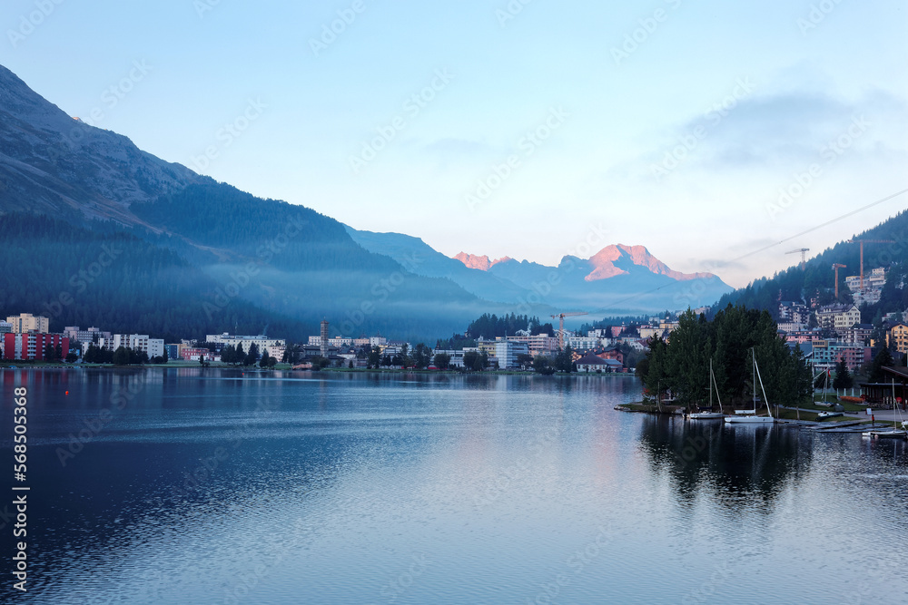 Morning scenery of beautiful Lake St. Moritz, a popular resort in Engadine, Switzerland, with sunrise glow on majestic Alpine mountains in background & village buildings by lakeside in misty twilight