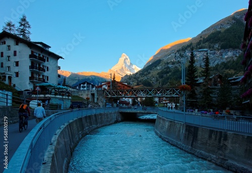 Morning scenery of Zermatt in twilight, a popular resort town in Valais, Switzerland, with view of tourists walking & cycling by a stream and majestic Mount Matterhorn brightened up by warm sunlight  © AaronPlayStation