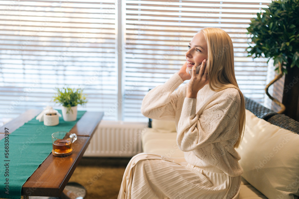Side view of pretty young woman smiling talking on mobile phone sitting on sofa at table with tea by window. Happy blonde female conversation with smartphone while rest indoors. Side view of pretty young woman smiling talking on mobile phone sitting on sofa at table with tea by window. Happy blonde female conversation with smartphone while rest indoors.