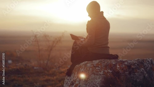  Young Christian woman girl reading the bible, praying, putting her hand on the bible, sitting on a rock in nature with an incredible view, sunset, peace and tranquility, faith. High quality 4k 