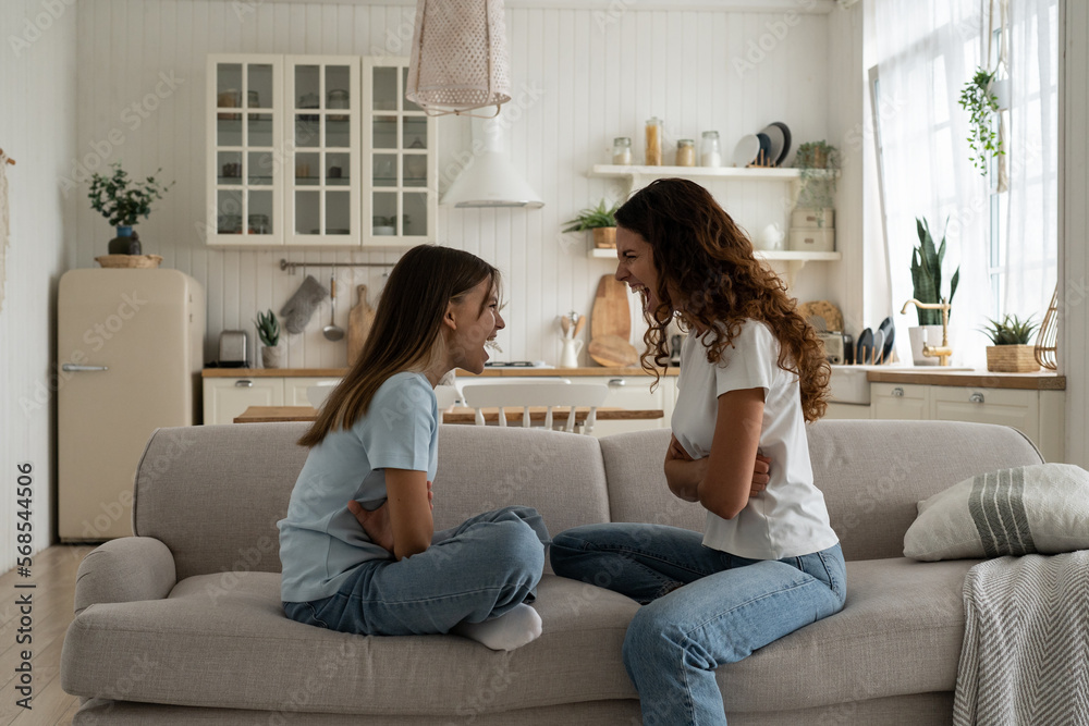 Cursing nervous woman and teen girl are sits on couch and shouting out loud expressing resentment and feeling mutual dislike. Aggressive mom and daughter arguing not wanting to give in to each other Cursing nervous woman and teen girl are sits on couch and shouting out loud expressing resentment and feeling mutual dislike. Aggressive mom and daughter arguing not wanting to give in to each other