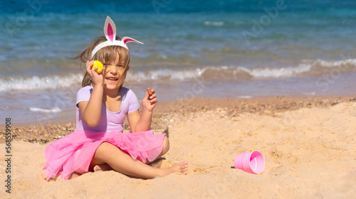 Happy 4 years old girl in a pink skirt sitting with colorful eggs on the sandy beach. Happy Easter holidays concept 