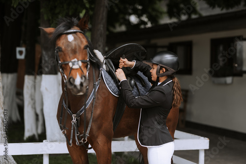 Girl rider adjusts saddle on her horse to take part in horse races. © JJ Studio