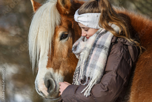 Cute equestrian and horse friendship scene: A young teenage girl interacting with her haflinger horse in front of a snowy rural landscape in winter outdoors