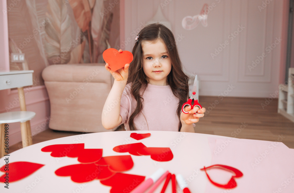 a little girl makes Valentine's Day cards using colored paper, scissors and pencil, sitting at a table in a cozy room. a little girl makes Valentine's Day cards using colored paper, scissors and pencil, sitting at a table in a cozy room.
