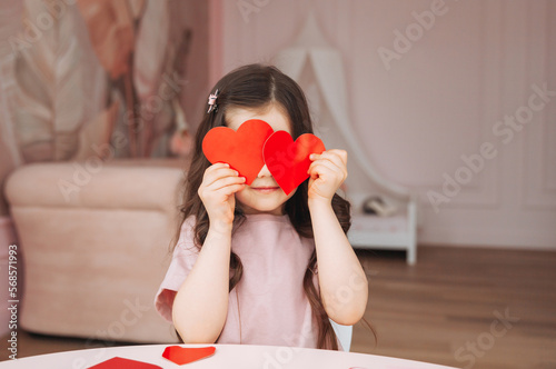 a little girl makes Valentine's Day cards using colored paper, scissors and pencil, sitting at a table in a cozy room. photo