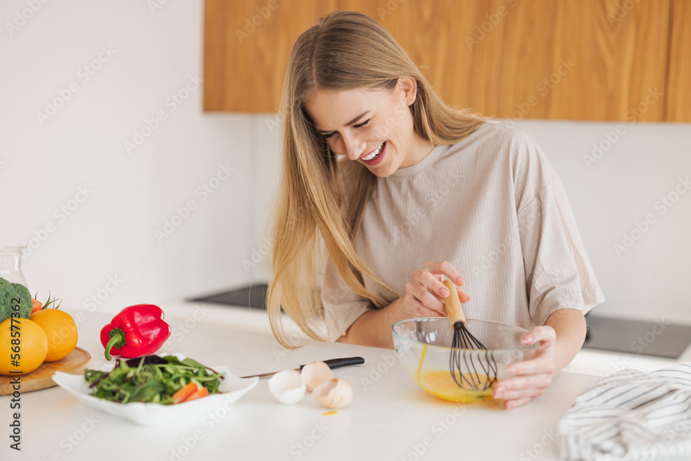 Happy pretty blonde woman in pajamas is beating eggs for breakfast in the kitchen at home.