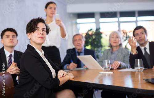 Office coworkers of different ages watching presentation while sitting at table with laptop and notes