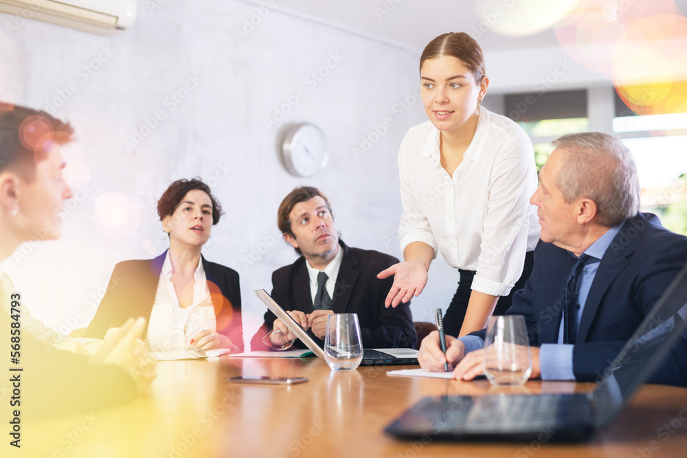 Positive young businesswoman discussing new business project with members of team gathered around table in office meeting room