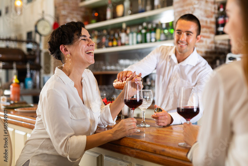 Two smiling female friends sitting in the bar and drinking red wine while bartender pouring some drink