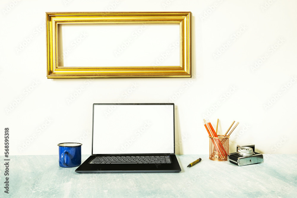 Fototapeta premium Image of a laptop on a blue surface next to a vintage camera, a coffee mug and a gold frame on the wall