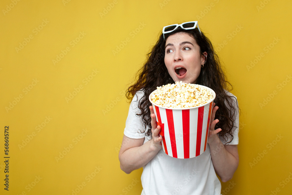 young shocked girl viewer in 3d glasses and a white t-shirt holds a large bucket of popcorn and shows surprise