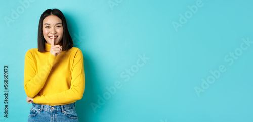 Cheerful asian girl asking to be queit, shushing at camera and smiling, keeping secret, standing against blue background photo