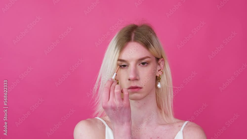 Young transgender man transsexual applying make-up on a pink background Young transgender man transsexual applying make-up on a pink background