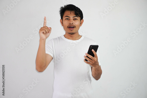 Excited young Asian man pointing fingers up having a good idea with holding mobile phone wearing white t shirt isolated on white background