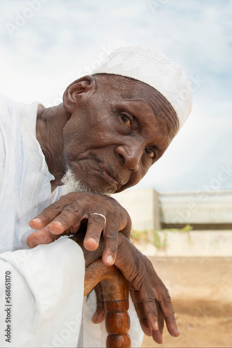 Old African man sitting in front of his house, looking at camera, serious expression, celebration clothing and hat, wooden, cane, eighty years old, photo
