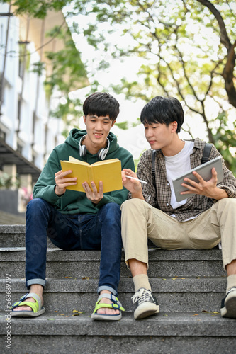 Two Asian male college students having break after classes outside of the building, reading book