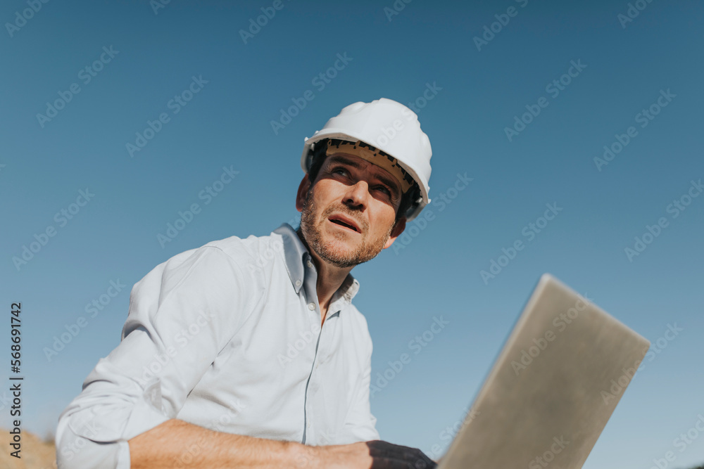 Thoughtful mature architect with laptop sitting in front of blue sky Thoughtful mature architect with laptop sitting in front of blue sky