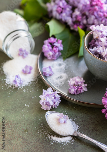 Flavored sugar with lilac flowers in glass jar and branch of beautiful lilac, green concrete background. Edible flowers in cooking and confectionery. Flavoring ingredients.