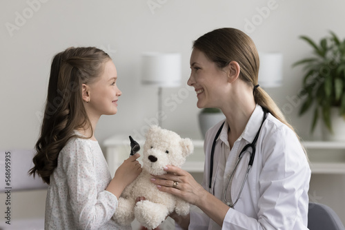 Fototapeta Naklejka Na Ścianę i Meble -  Happy pretty female pediatrician playing with little patient girl, using medical examination tools and teddy bear toy, chatting with kid, laughing, getting trust for checkup