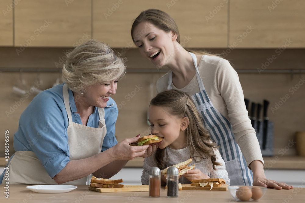Cheerful little girl, mom and grandma enjoying culinary activity in kitchen, cooking fast food snacks for lunch. Happy grandmother giving sandwich for kid biting, taste, feeding hungry child Cheerful little girl, mom and grandma enjoying culinary activity in kitchen, cooking fast food snacks for lunch. Happy grandmother giving sandwich for kid biting, taste, feeding hungry child