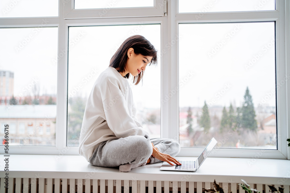 Smiling woman using laptop on window sill at home Smiling woman using laptop on window sill at home